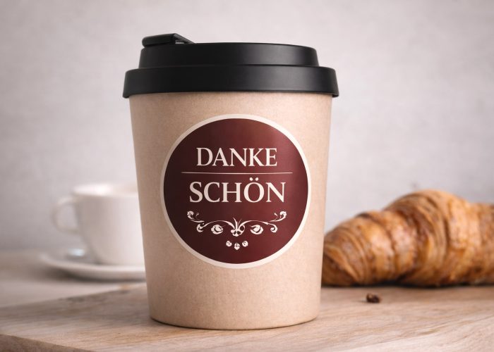 Kaffeebecher mit elegantem Danke-Schön Sticker auf Holztisch mit Croissant im Hintergrund